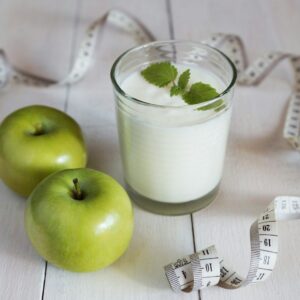 a glass of milk next to two green apples and a measuring tape
